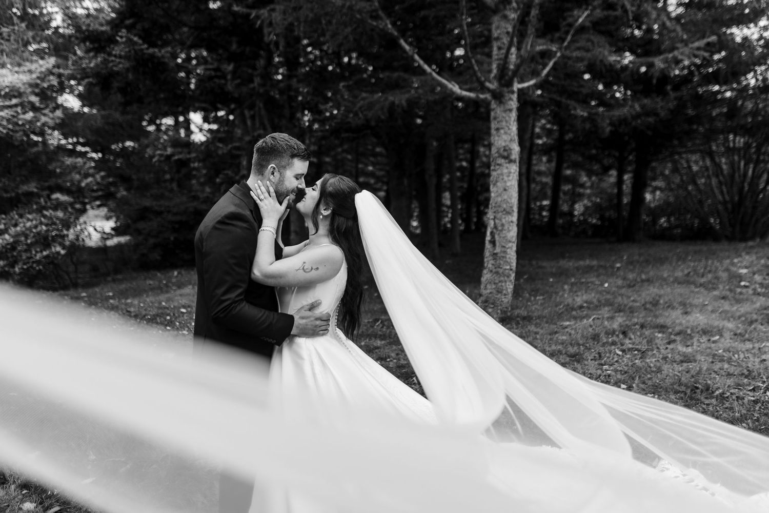 Black and white photo of a bride and groom sharing a kiss outdoors among trees, with her long wedding veil flowing in the foreground.