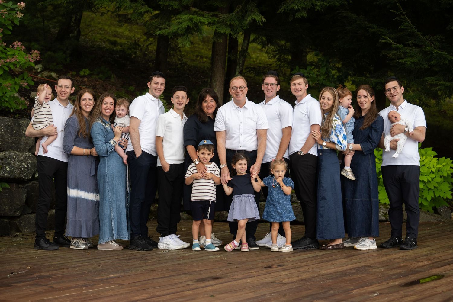 Large multigenerational family group portrait outdoors on wooden deck surrounded by lush green trees.