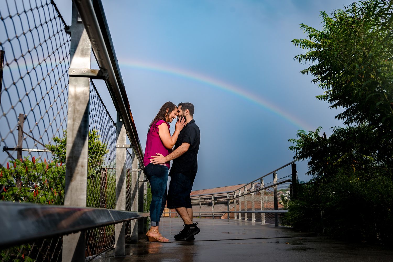 Romantic couple embracing on a bridge with a beautiful rainbow in the background during sunset.