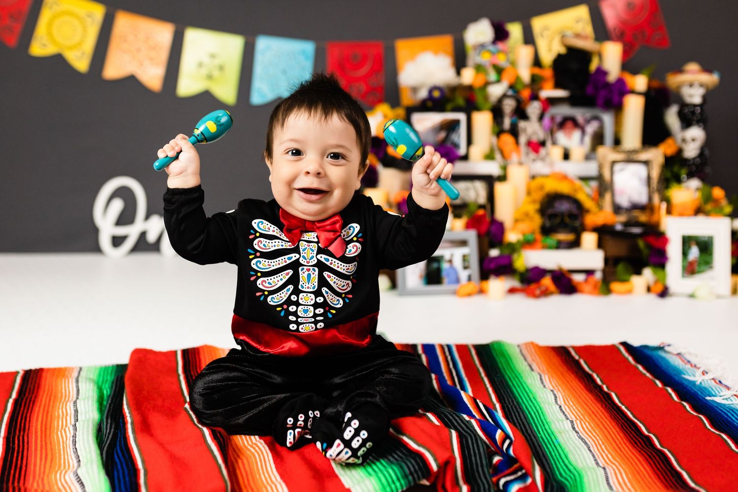 Small child in skeleton costume sitting on colorful striped blanket with festive decorations in background.