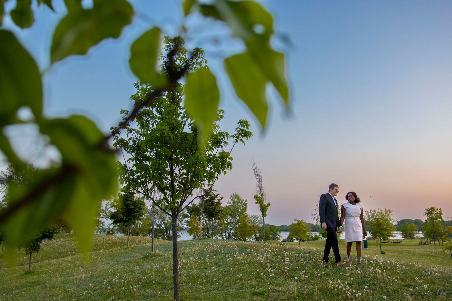 Two silhouettes stand in a field at sunset with young trees in the background.