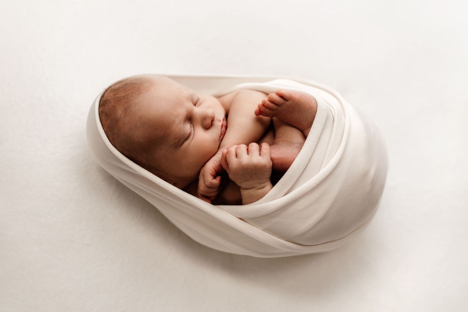 Peaceful newborn wrapped in soft white fabric against neutral background.