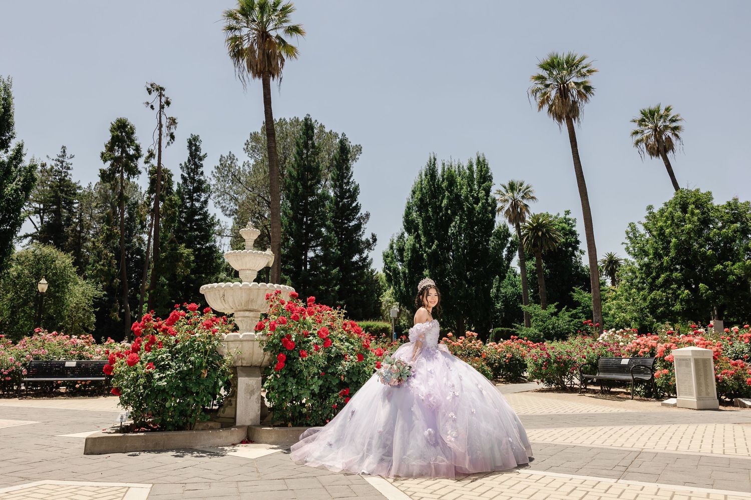 A lavender ballgown stands out against palm trees and red flowers in a sunlit garden setting.