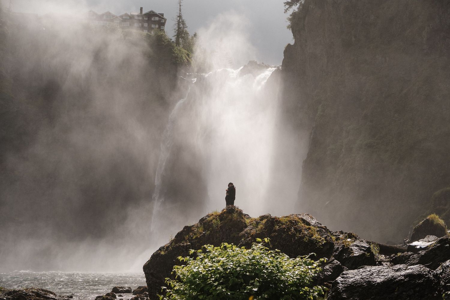 Lone figure standing on mossy rocks before a misty cascading waterfall in nature.