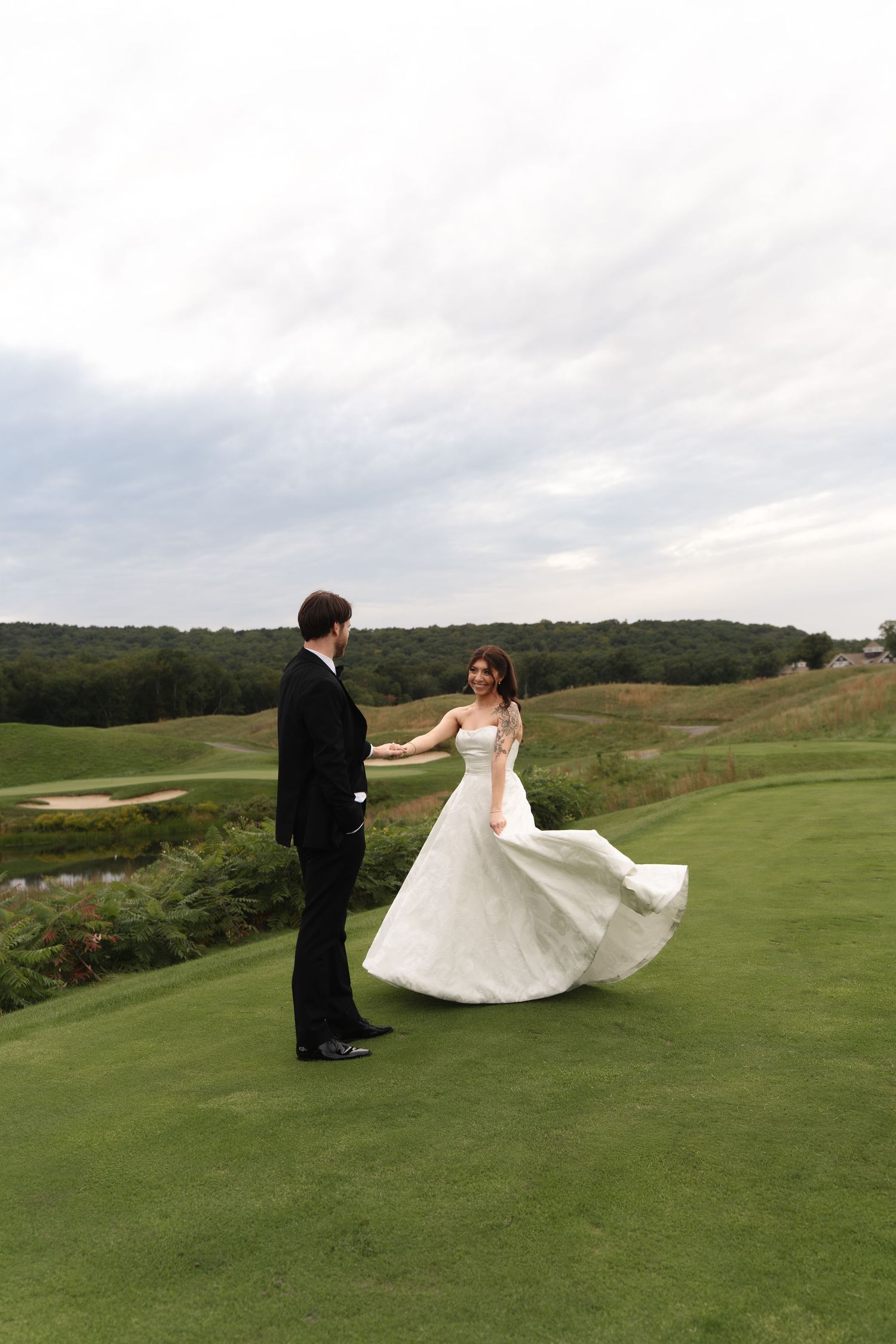 Bride in white gown twirls with groom in black tuxedo on lush green golf course under cloudy sky.