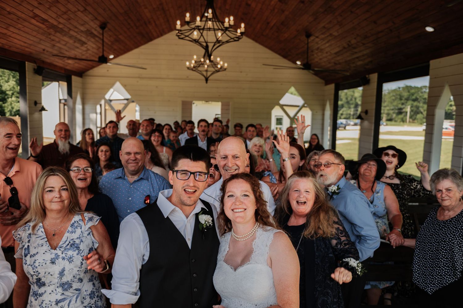 Bride and groom exchange vows at rustic indoor wedding ceremony with white chairs and floral arrangements.