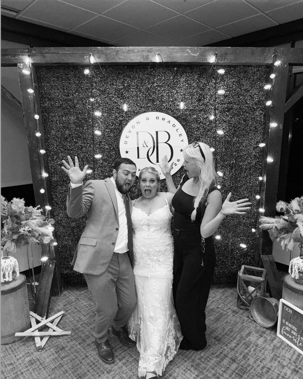 Black and white photo of bride in wedding dress posing with two guests in front of floral backdrop with string lights.