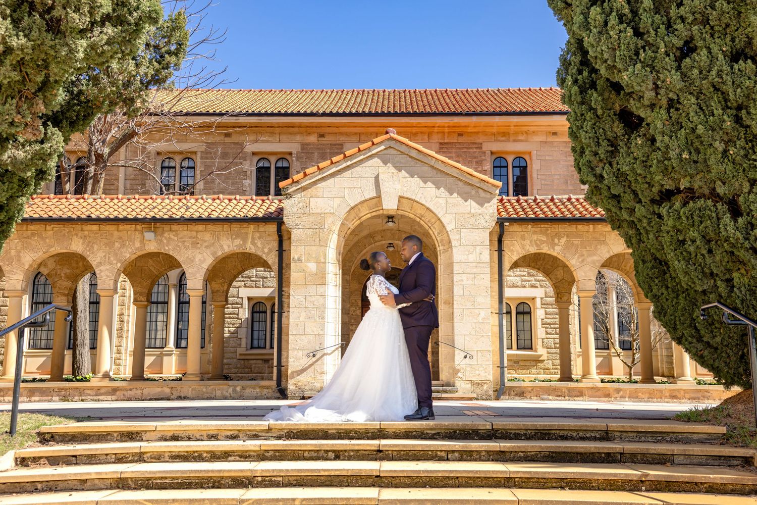 Bride and groom share romantic moment on steps of elegant stone building with arched colonnade on sunny wedding day.