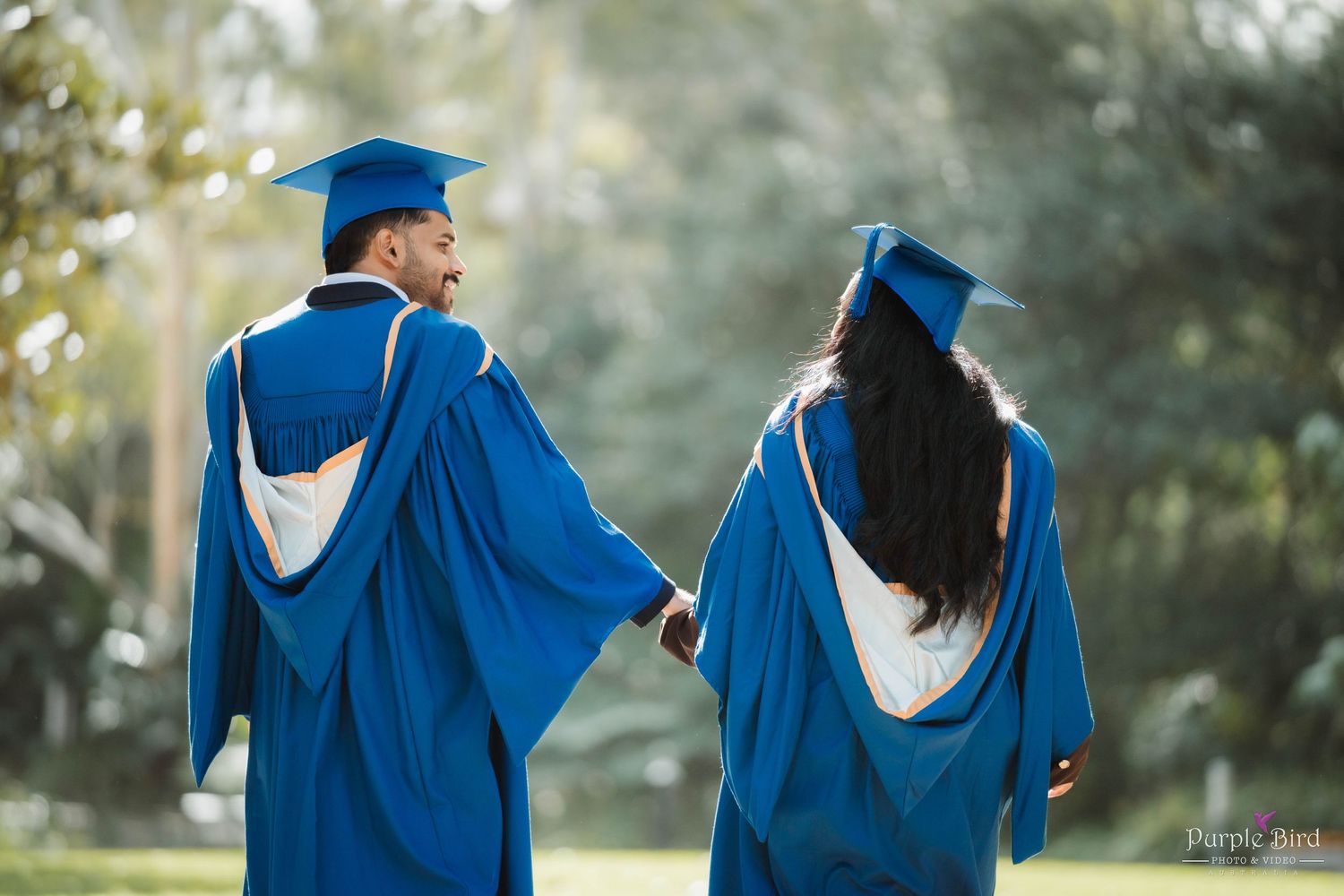 Two graduates in blue caps and gowns walk hand in hand across a sunlit green park, seen from behind, celebrating graduation day.
