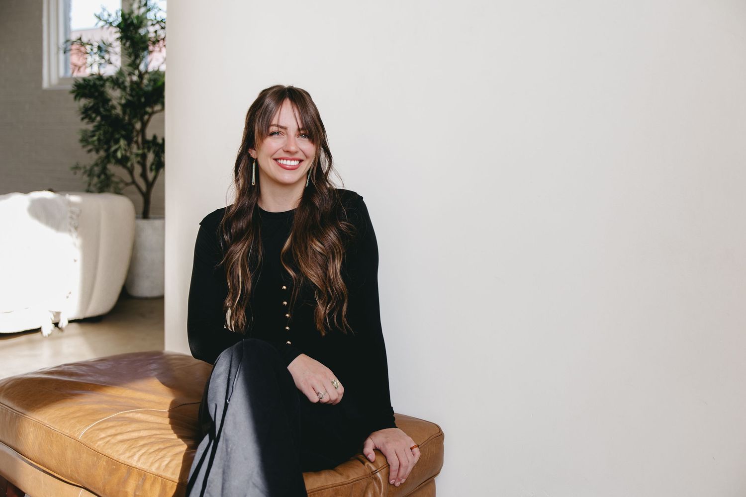 woman sitting on a leather foot rest against a white wall with a white couch in the background
