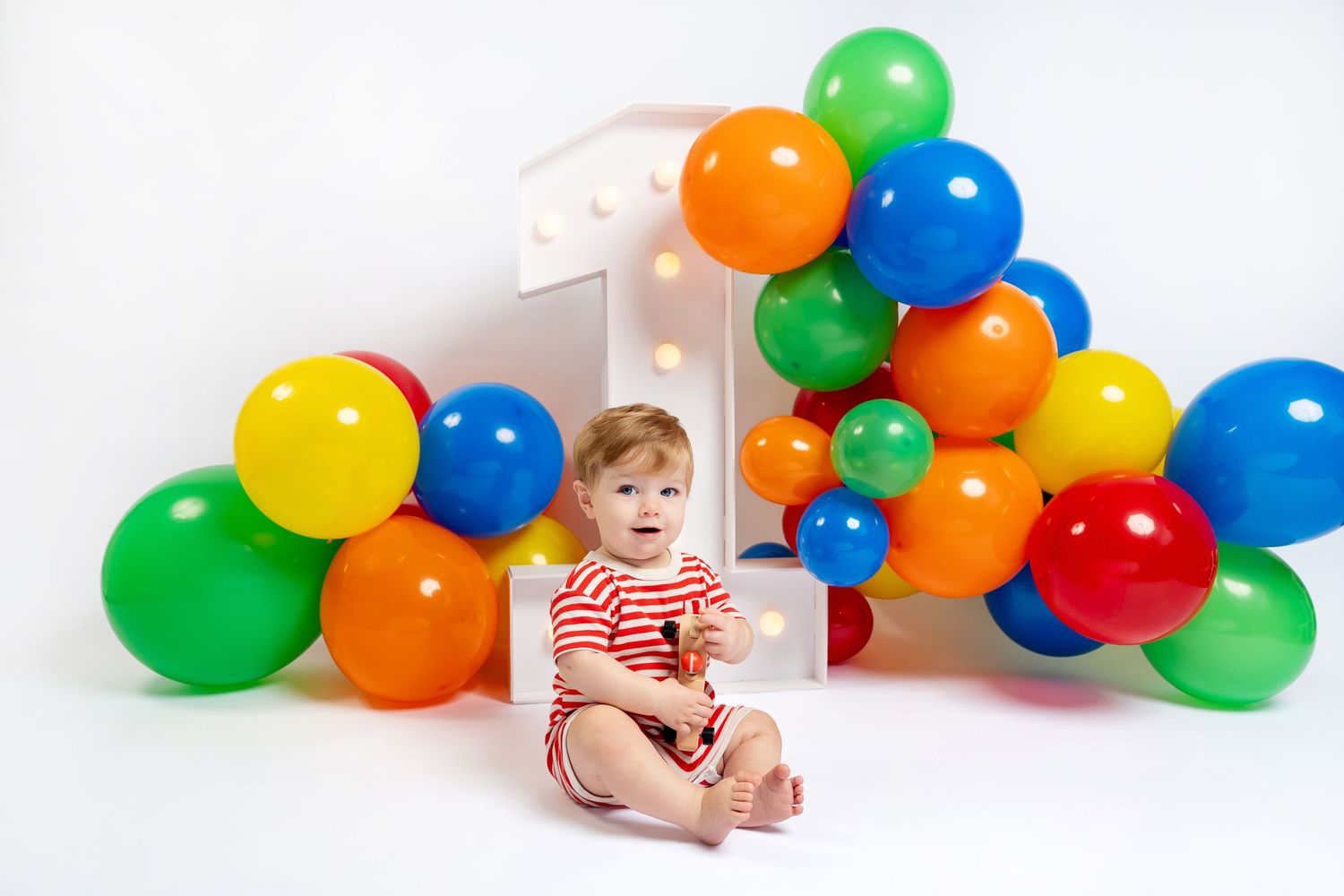 Baby's first birthday photo with colorful balloons and a large number one on a white background.