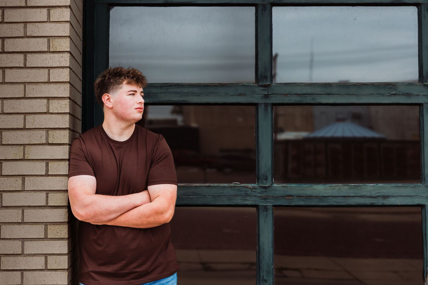 Young man in brown t-shirt stands arms crossed against brick wall near large window, looking away thoughtfully.
