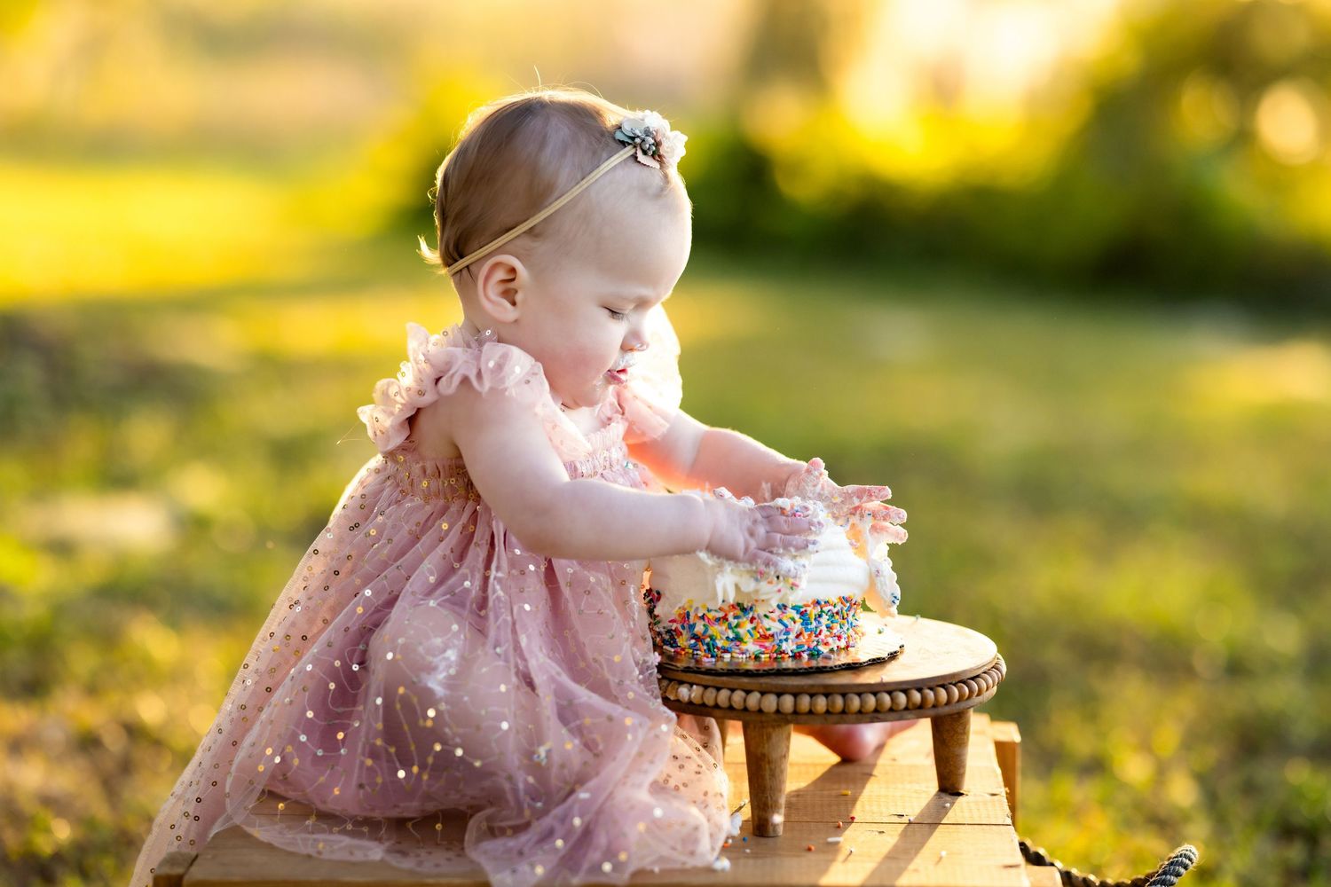 Baby girl in pink dress smashing her first birthday cake outdoors in golden sunlight.