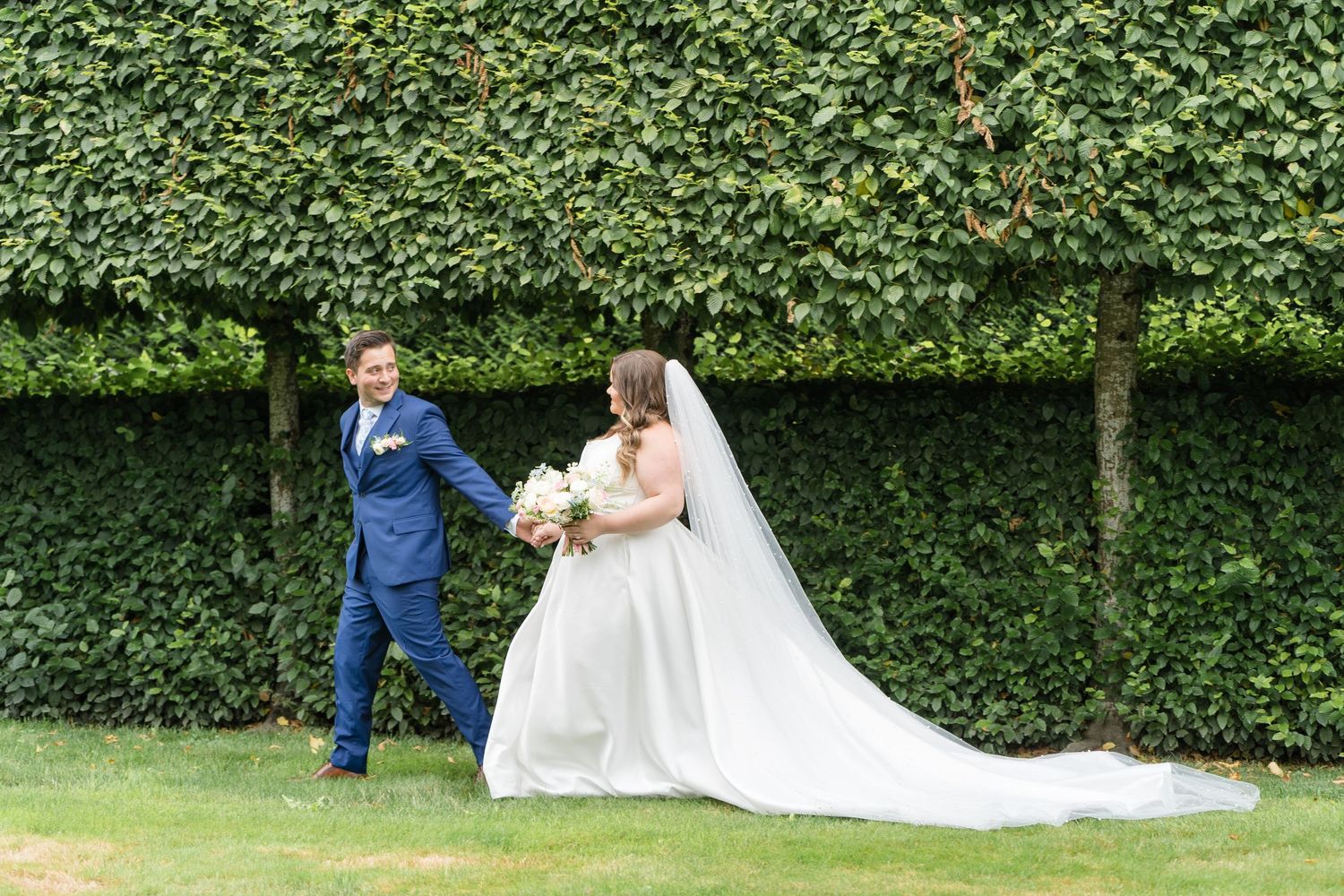 Bride and groom walk hand-in-hand on green lawn before lush ivy hedge wall on their wedding day.