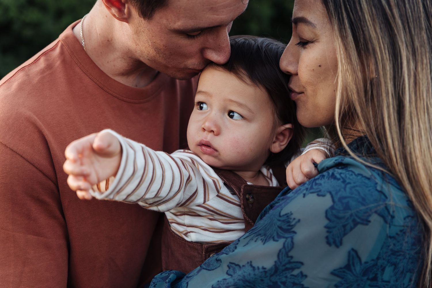 Parents kissing their curious baby boy on the head while the child points at something outdoors.