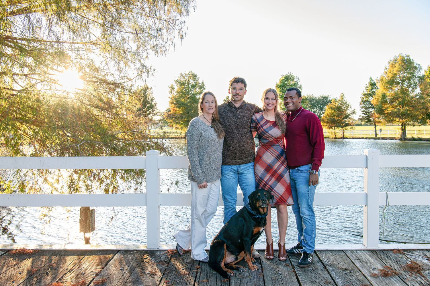 Four people and a Rottweiler pose on a wooden dock by a sunlit lake surrounded by autumn trees.