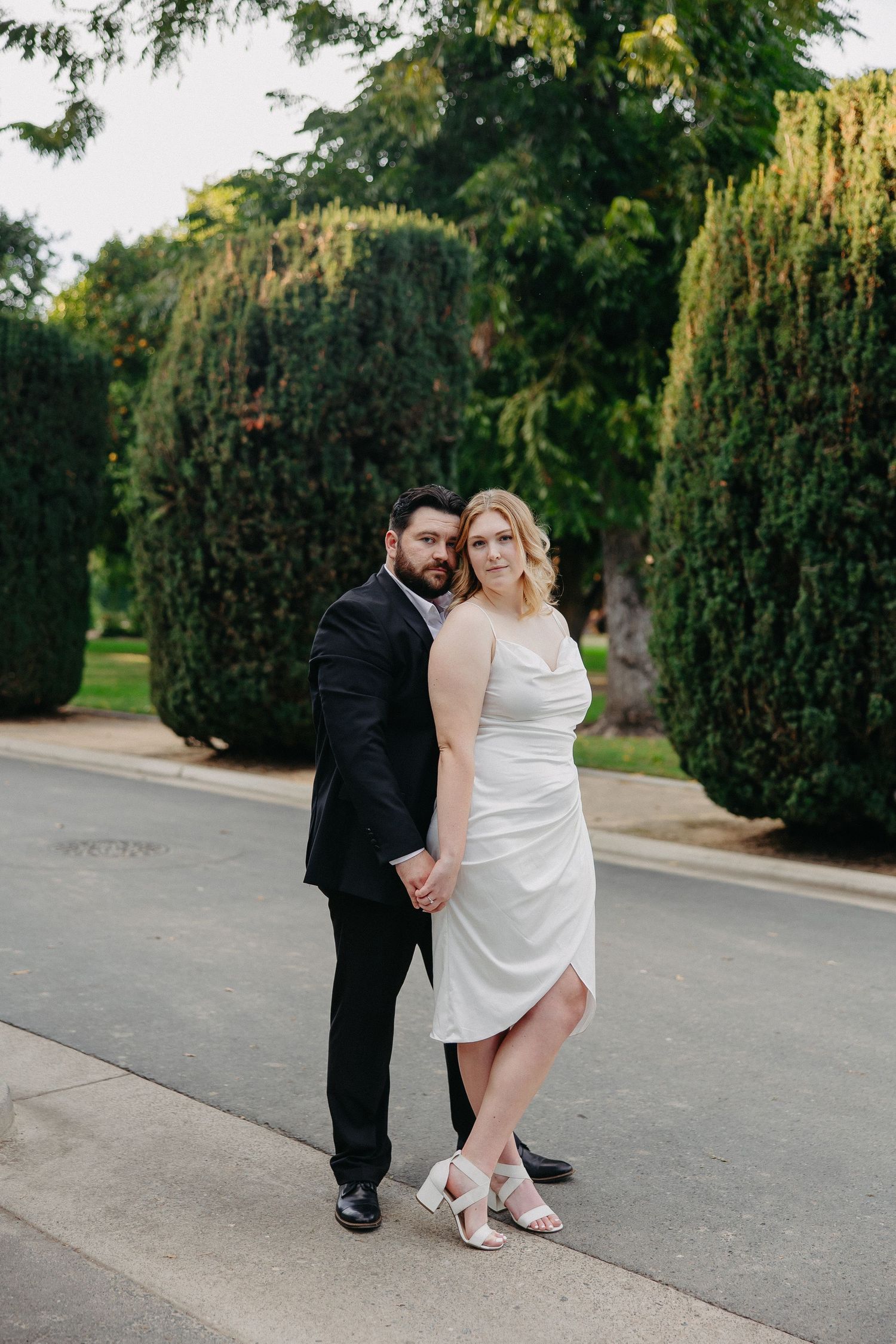 A couple poses outdoors in formal attire against lush green landscaping.