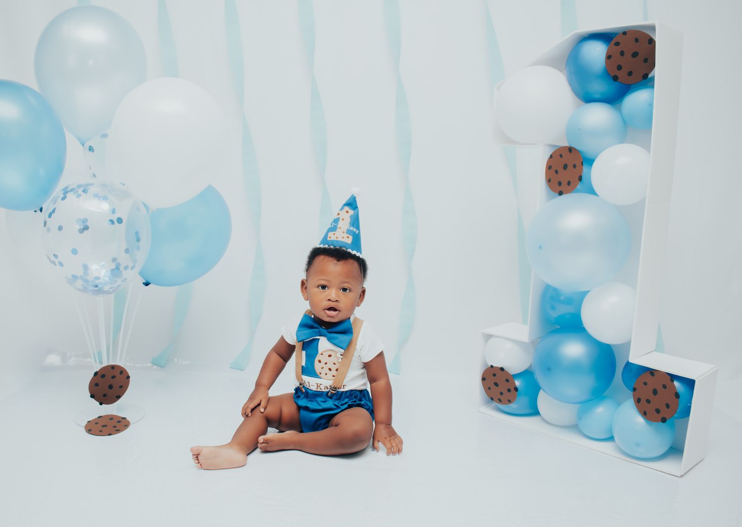 First birthday celebration setup with blue and white balloons and teddy bear decorations on white backdrop.