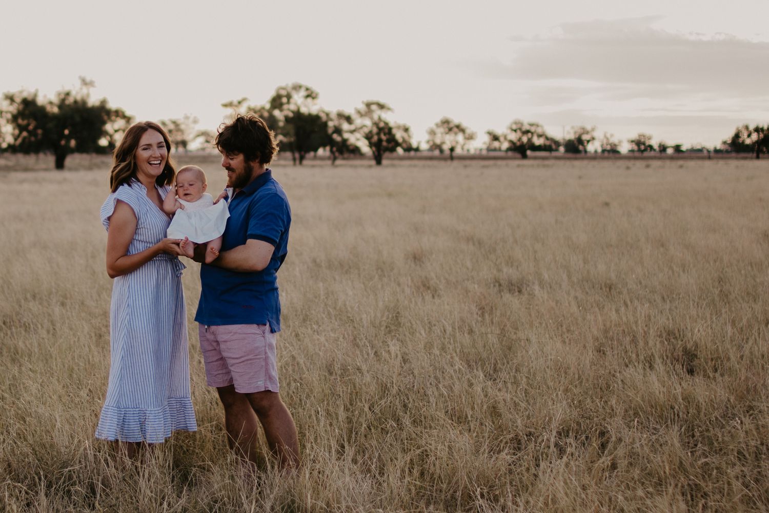 Mum & Dad holding baby girl between, in the paddock on their farm during golden hour at Armatree NSW.
