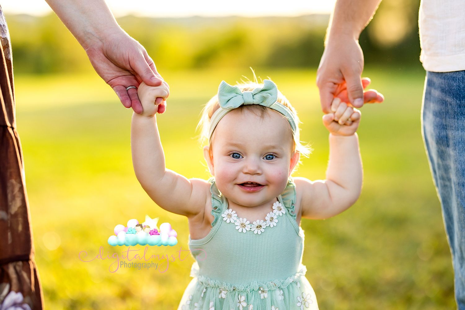 A smiling baby in a mint green dress and headband holds hands with adults while standing in a sunny field.