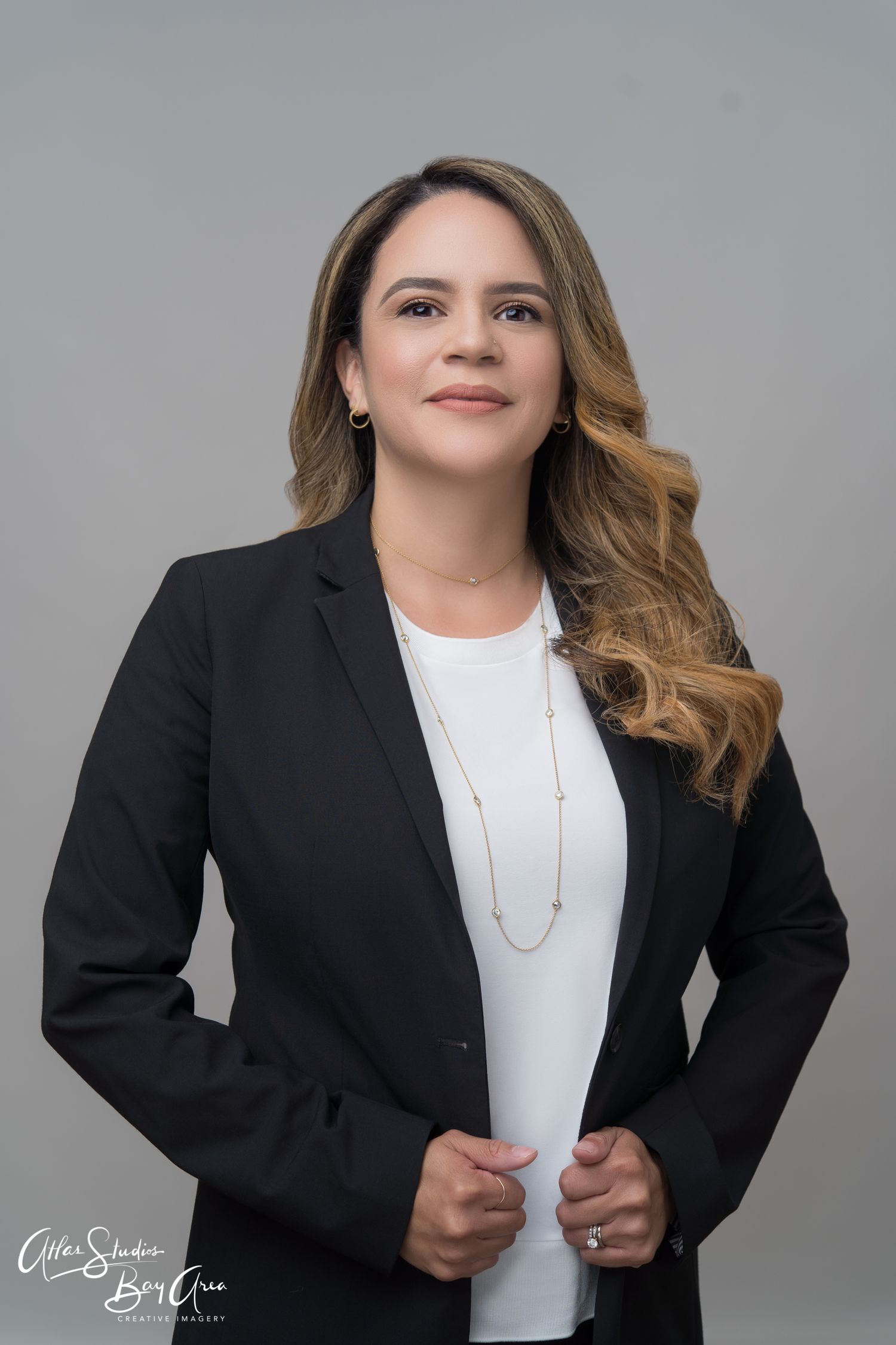 Professional headshot of a person in a black blazer and white top with flowing hair against a gray background.