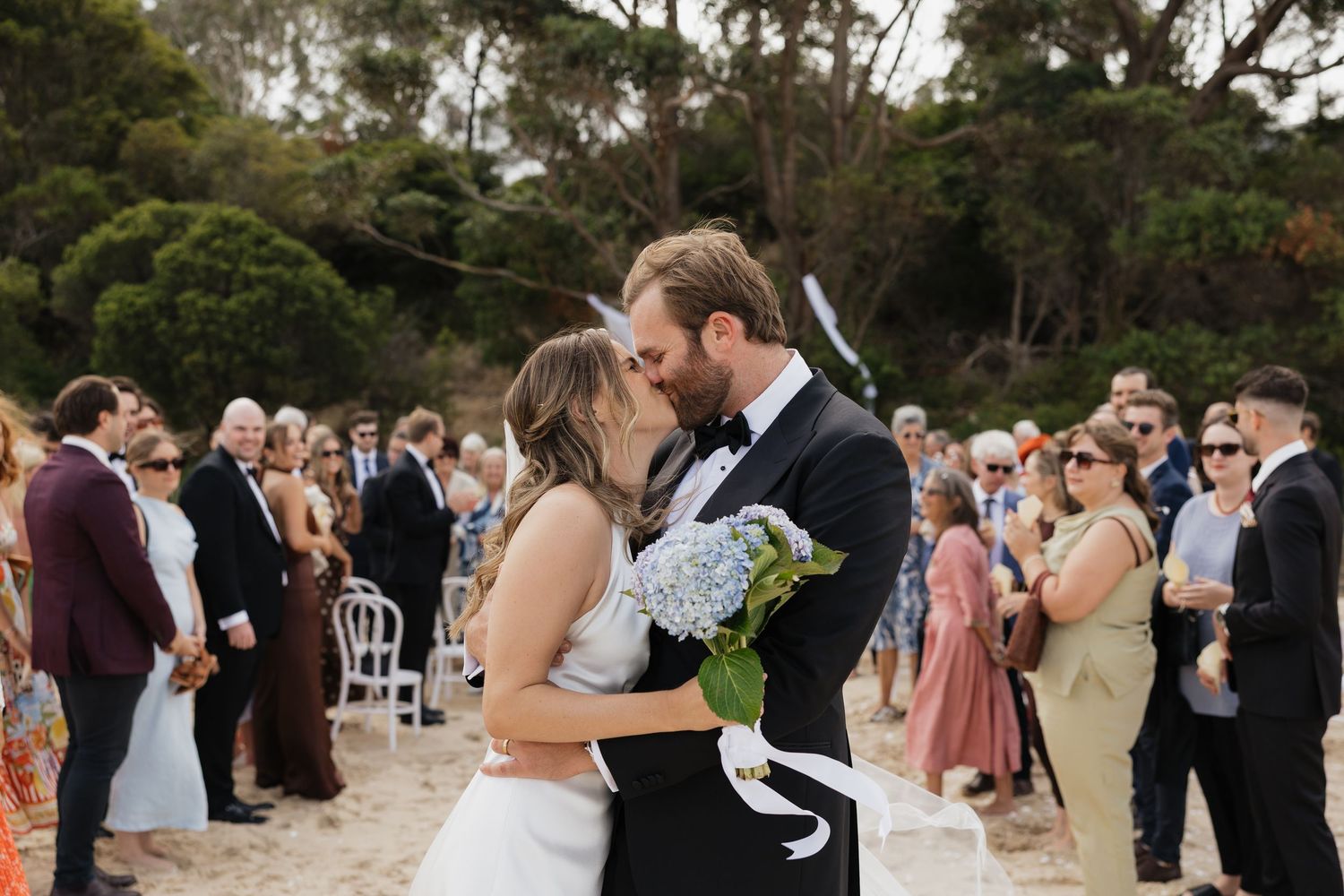 Wedding guests dance and celebrate together at an outdoor reception venue surrounded by trees.