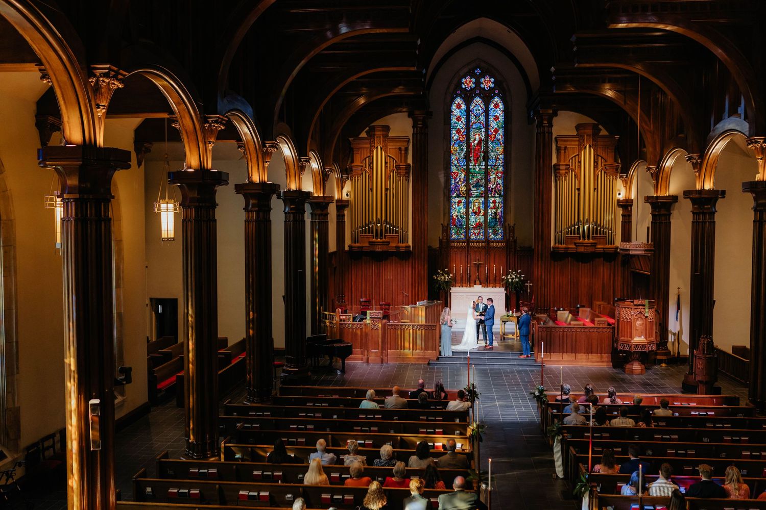 Interior view of a church sanctuary with gothic arches, stained glass windows and red altar area during a ceremony.