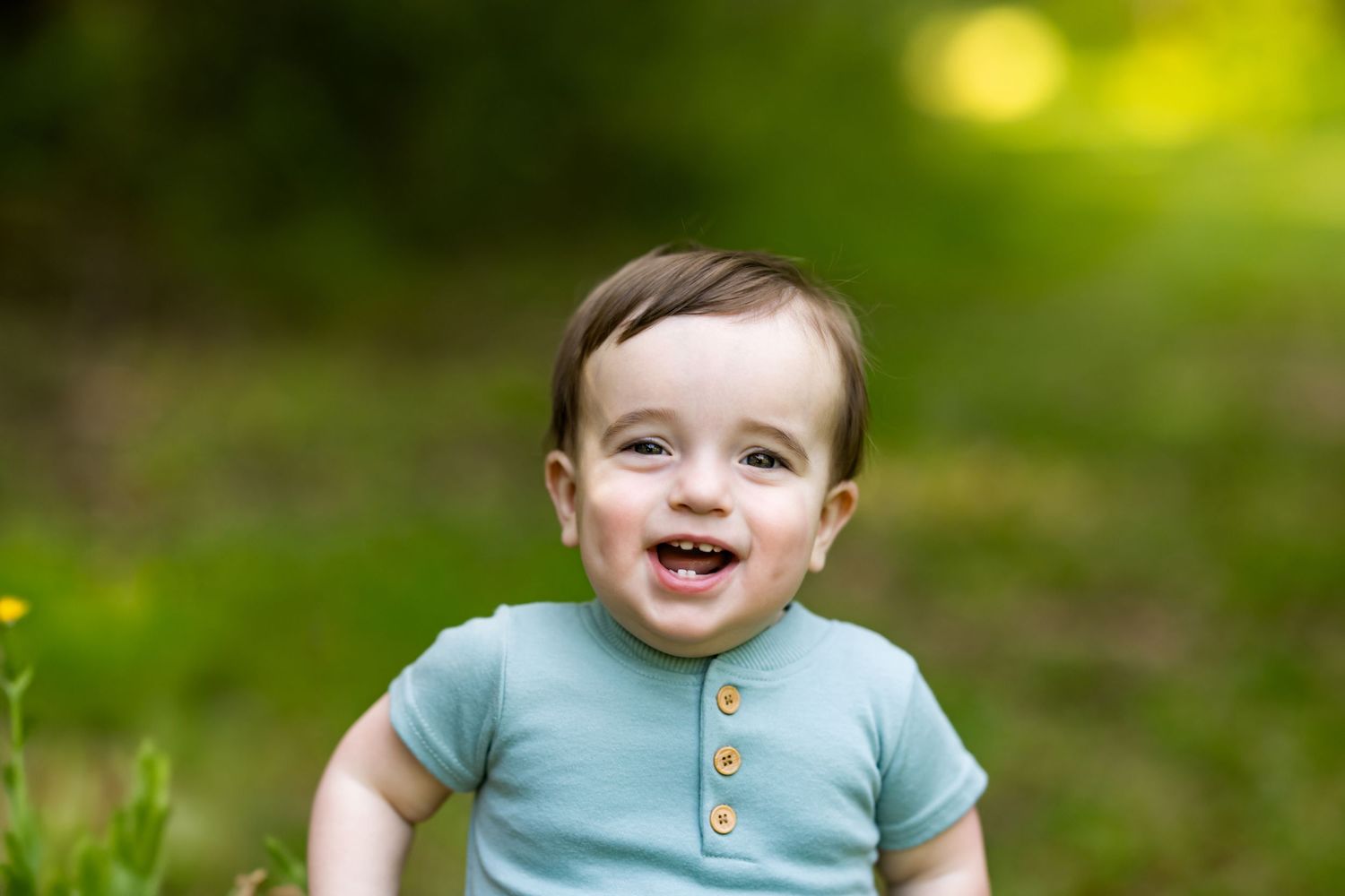 Young child sits in grass wearing teal shirt and laughing during outdoor portrait session in natural light.