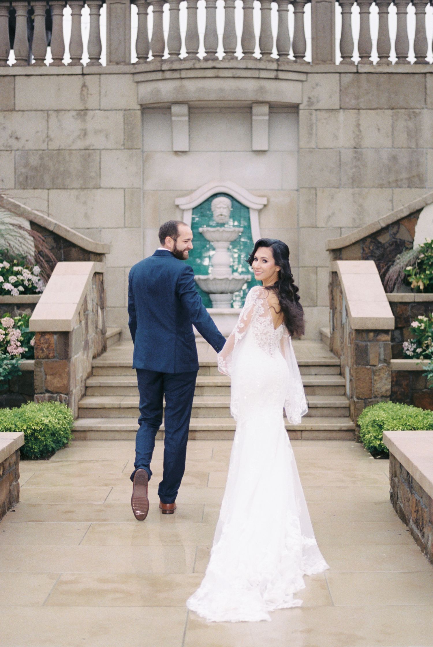 A wedding couple ascends stone steps in a classical architectural setting with white balusters and floral arrangements.