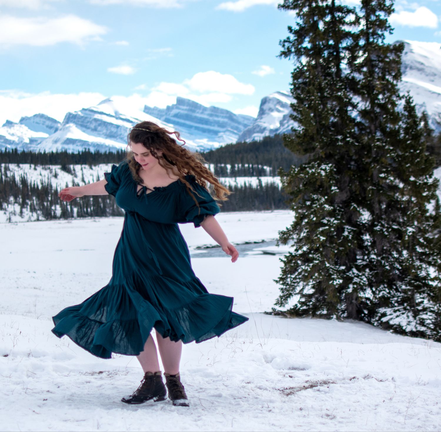A figure in a flowing navy dress twirls joyfully in the snowy mountain landscape with snow-capped peaks in the background.