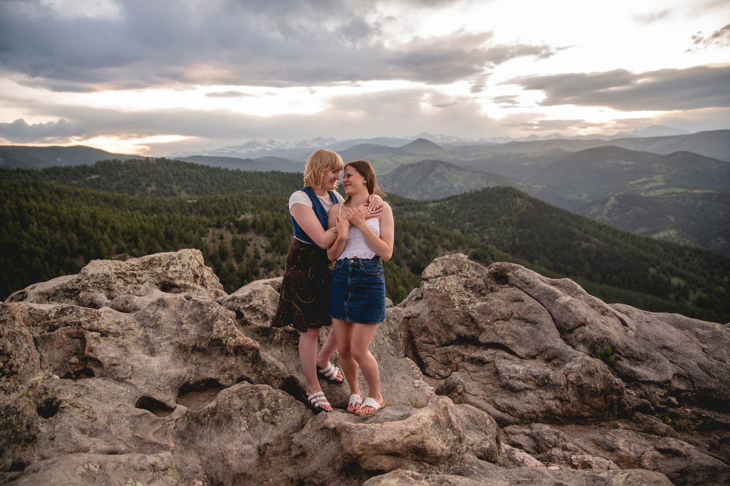 Mountain engagement photo session at sunset showing couple on rocky overlook with dramatic sky and mountain views in background.