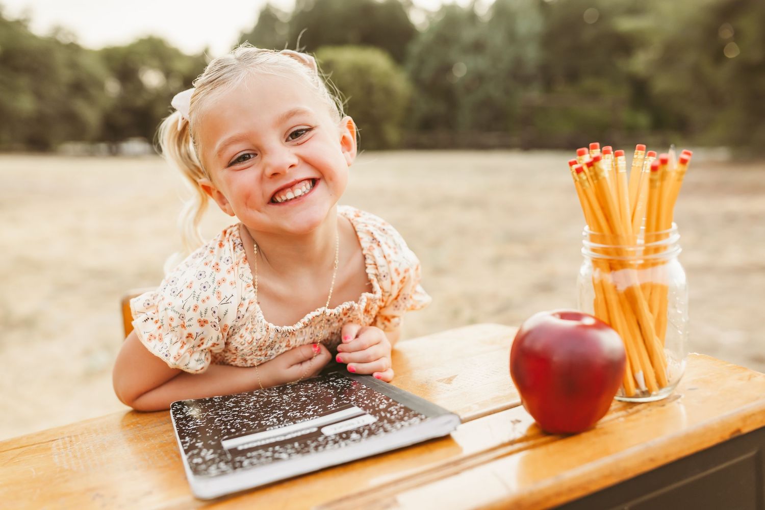 A student works on a composition notebook at a wooden desk with school supplies and a red apple.