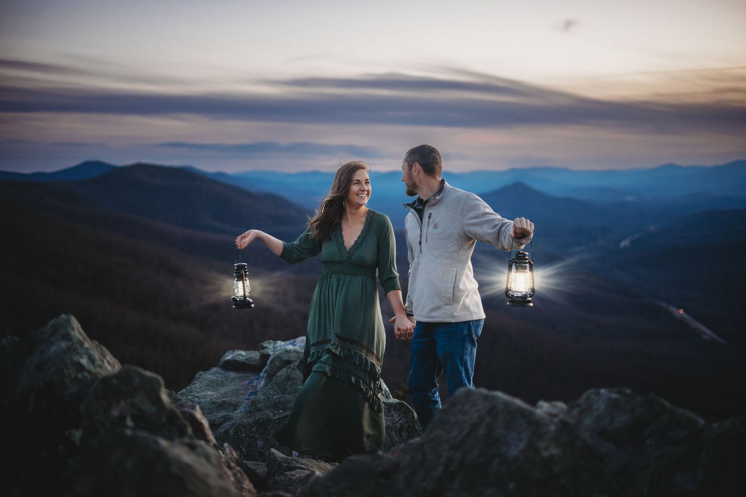 A couple enjoys a romantic sunset moment on a mountain overlook with dramatic blue ridge views in the background.