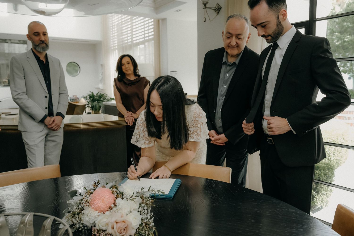 Wedding guests gather around a table during a reception, dressed in formal black attire.