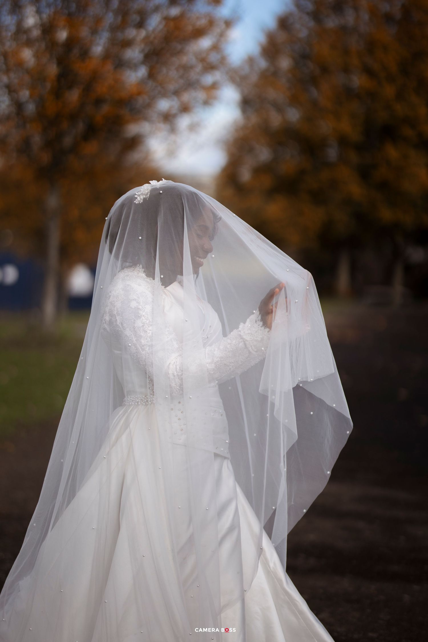 A bride in a flowing white wedding dress and veil poses outdoors during autumn with orange trees in the background.