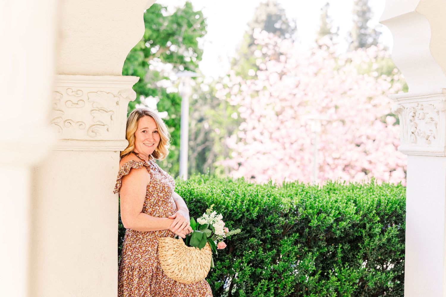 Woman in floral dress holding wicker bag stands by white columns with cherry blossoms and greenery in background.