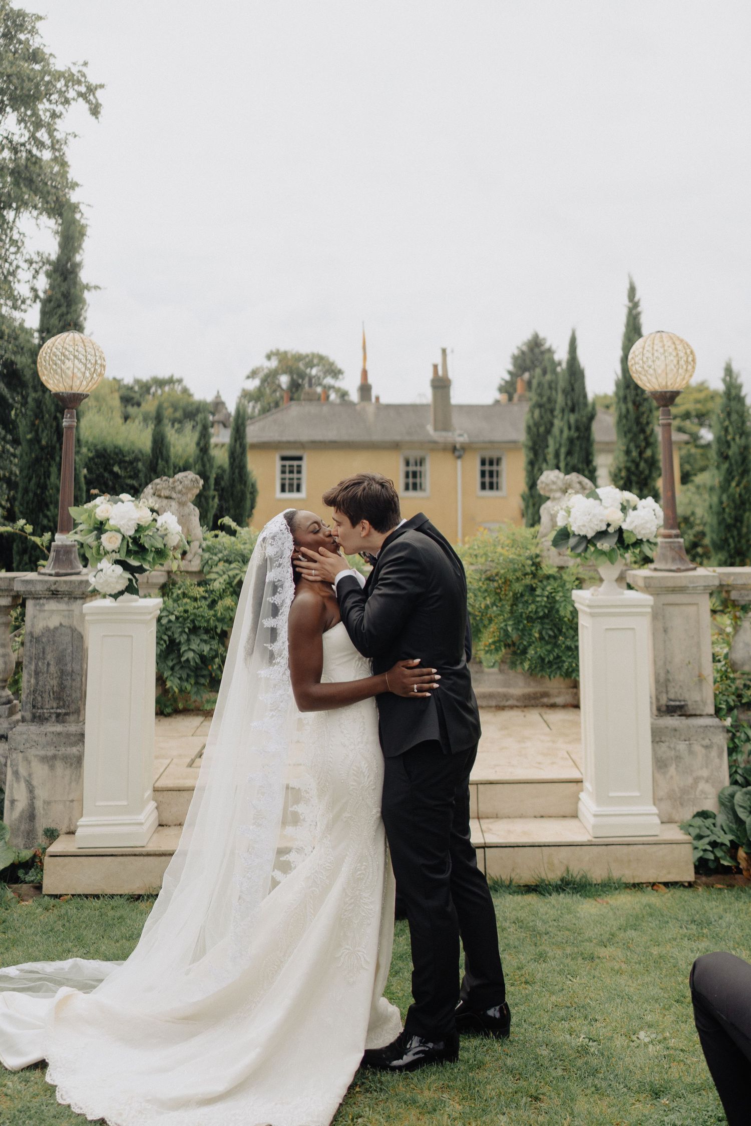 A romantic wedding ceremony moment in a garden setting with white floral arrangements and stone pillars.