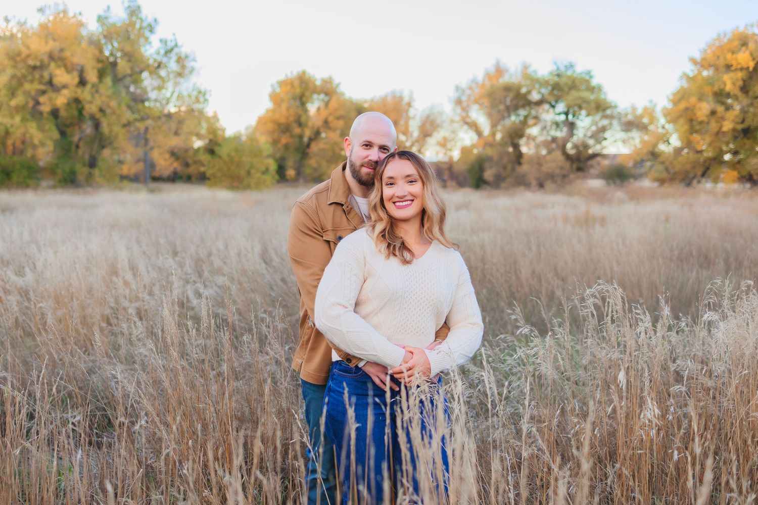 Couple embracing in golden autumn field with colorful trees, woman in cream sweater and man in brown shirt.  Man hugging woman from behind