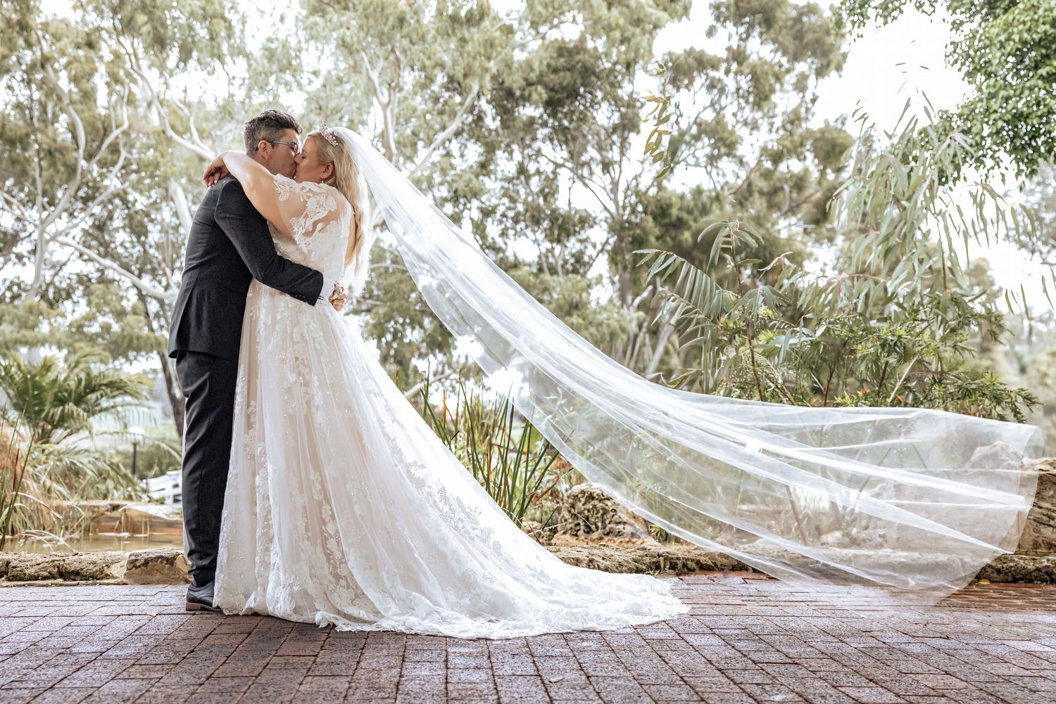 A romantic wedding couple embraces on a brick pathway surrounded by lush greenery and flowing white veil.