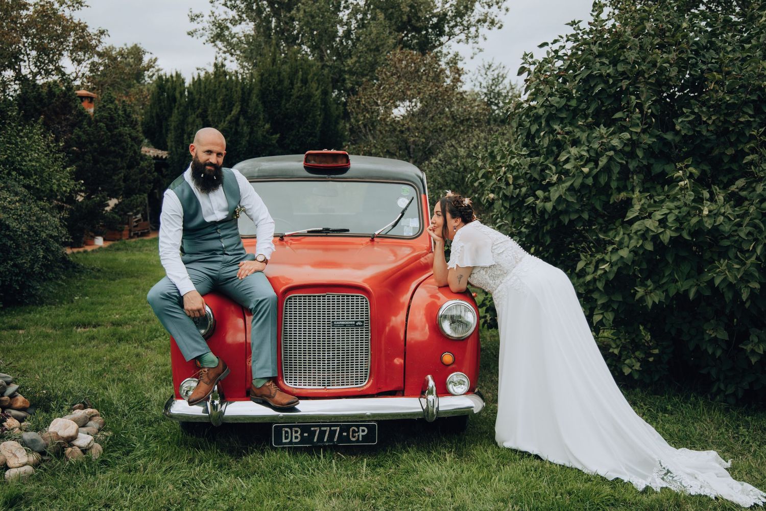 Bride and groom posing with vintage red car in lush garden setting on wedding day.