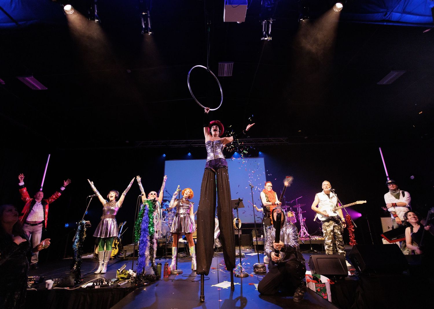 Colorful performance troupe on stage with stilt walker holding hoop under dramatic stage lighting.