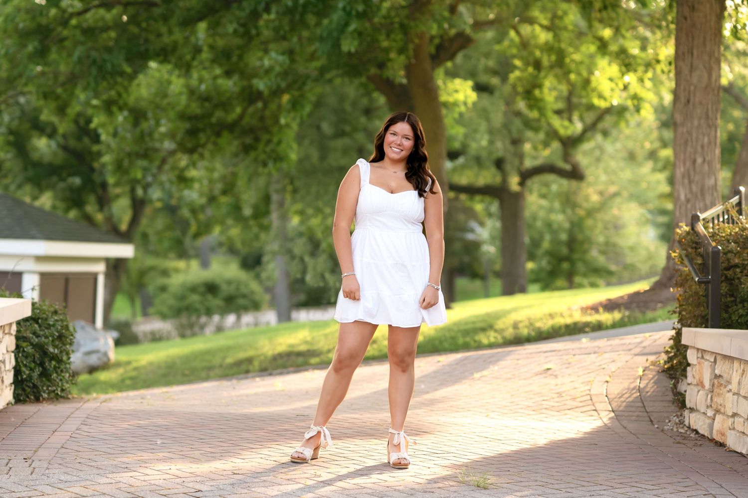 A person in a white dress walks along a sunlit stone path through a lush green garden setting.