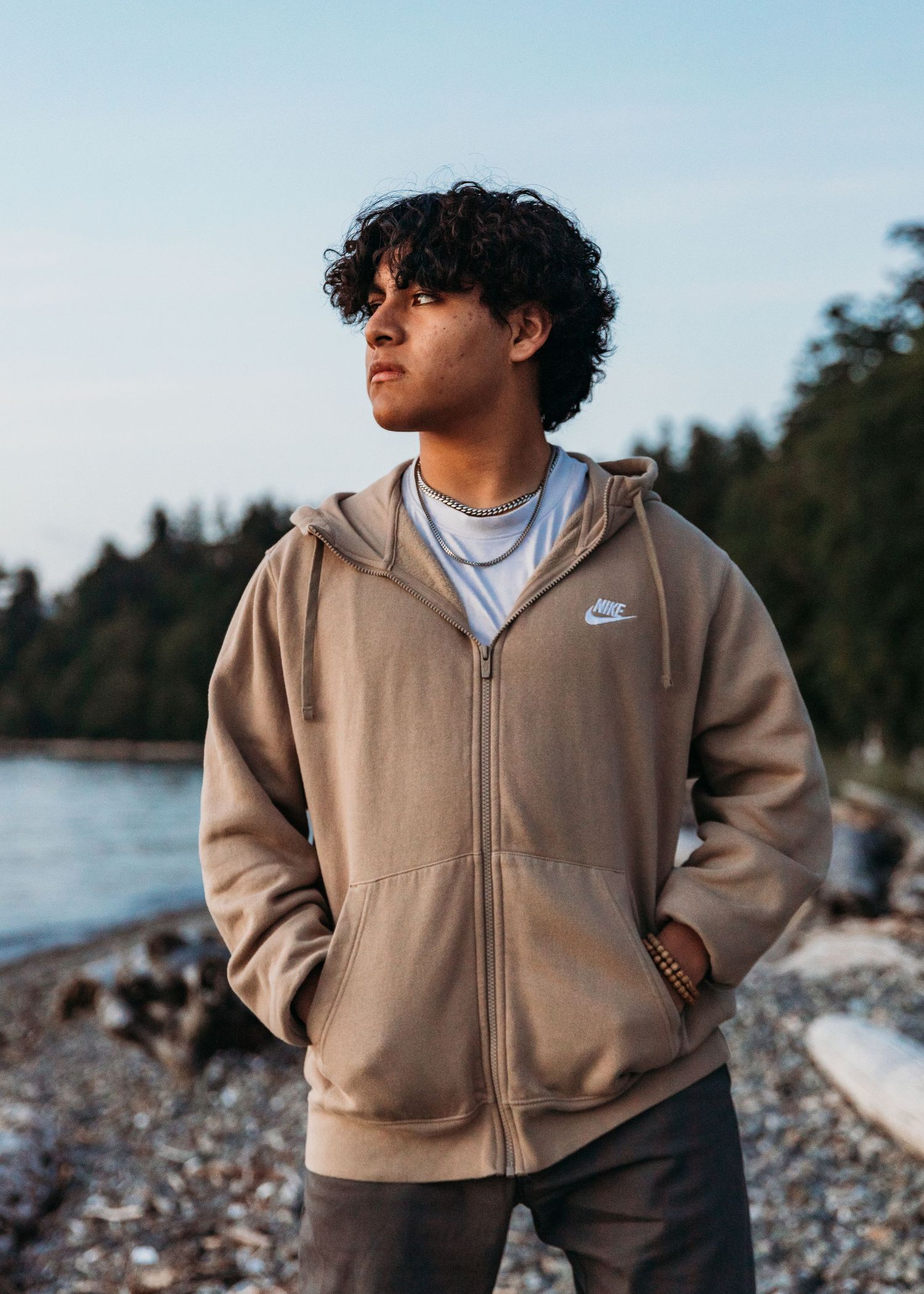Person wearing a tan Nike zip-up hoodie stands on a rocky beach shore at sunset with forest in background.