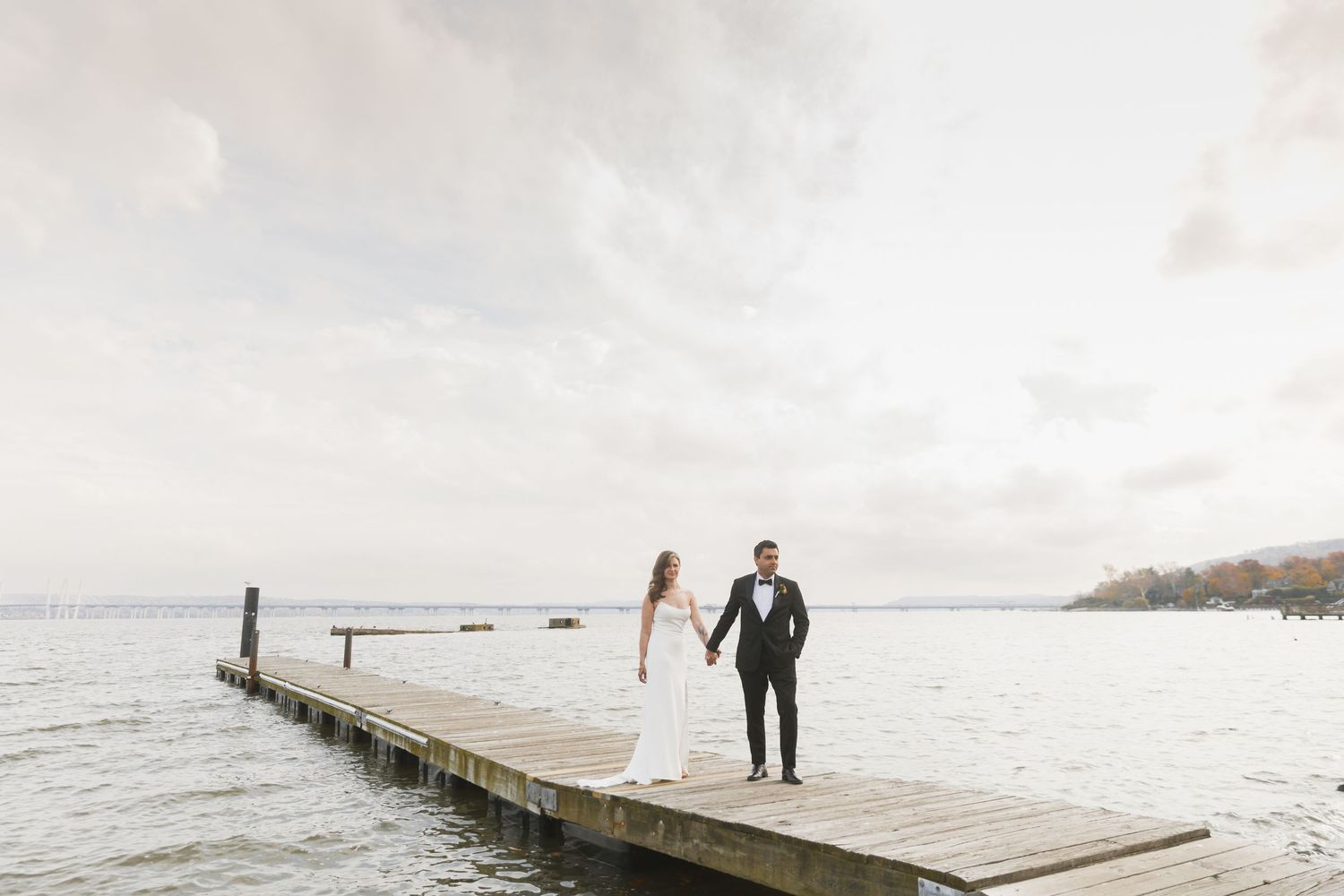 A couple in wedding attire walks together on a wooden dock extending into a serene lake on an overcast day.