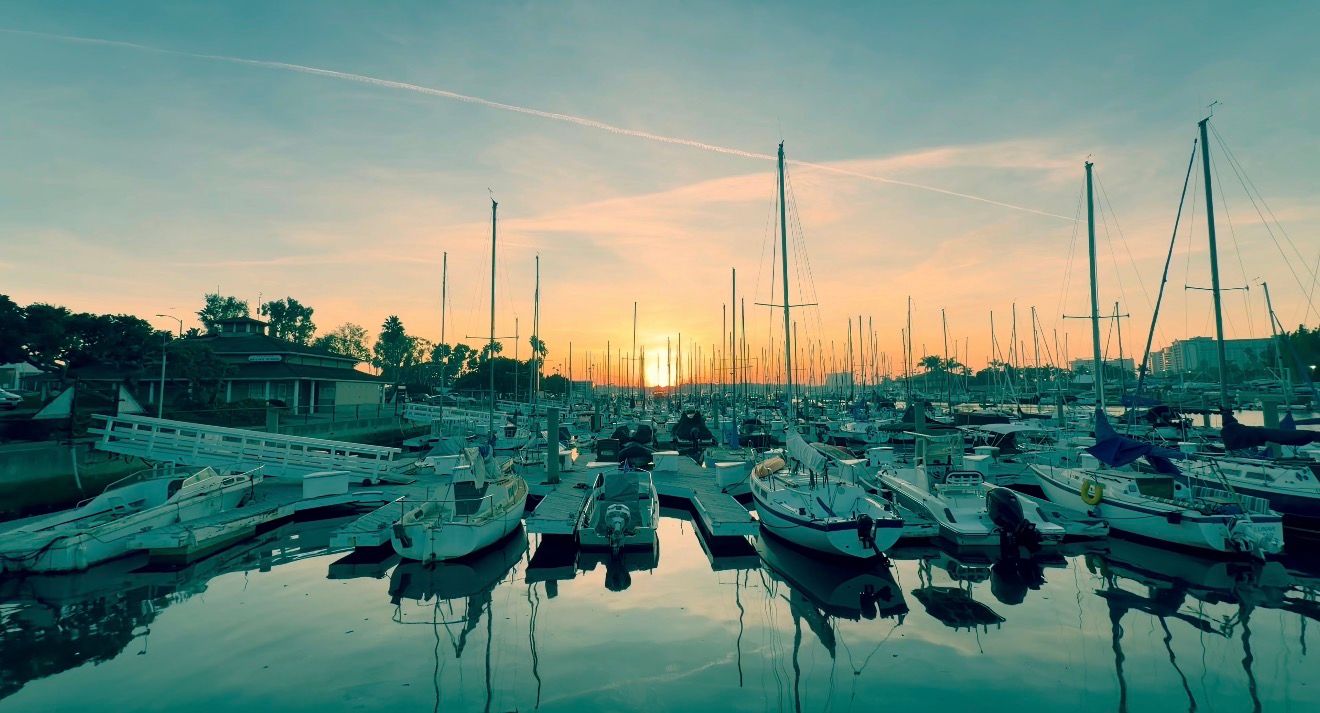 Panoramic view of sailboats moored in a marina at sunset with calm waters reflecting the colorful sky.