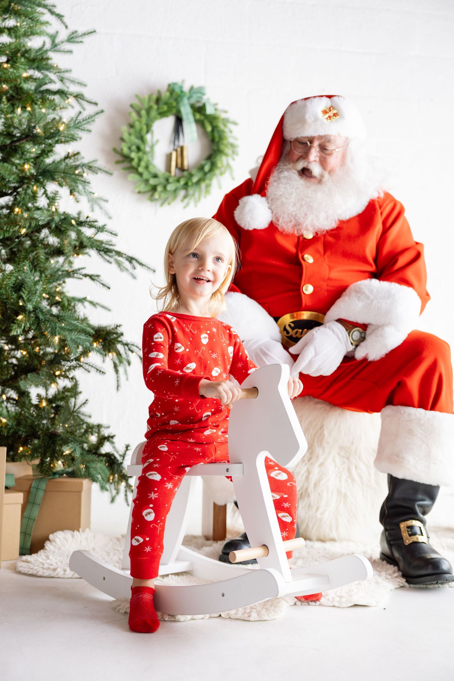 A child in red holiday pajamas plays with a rocking horse toy next to a decorated Christmas tree and festive decor.