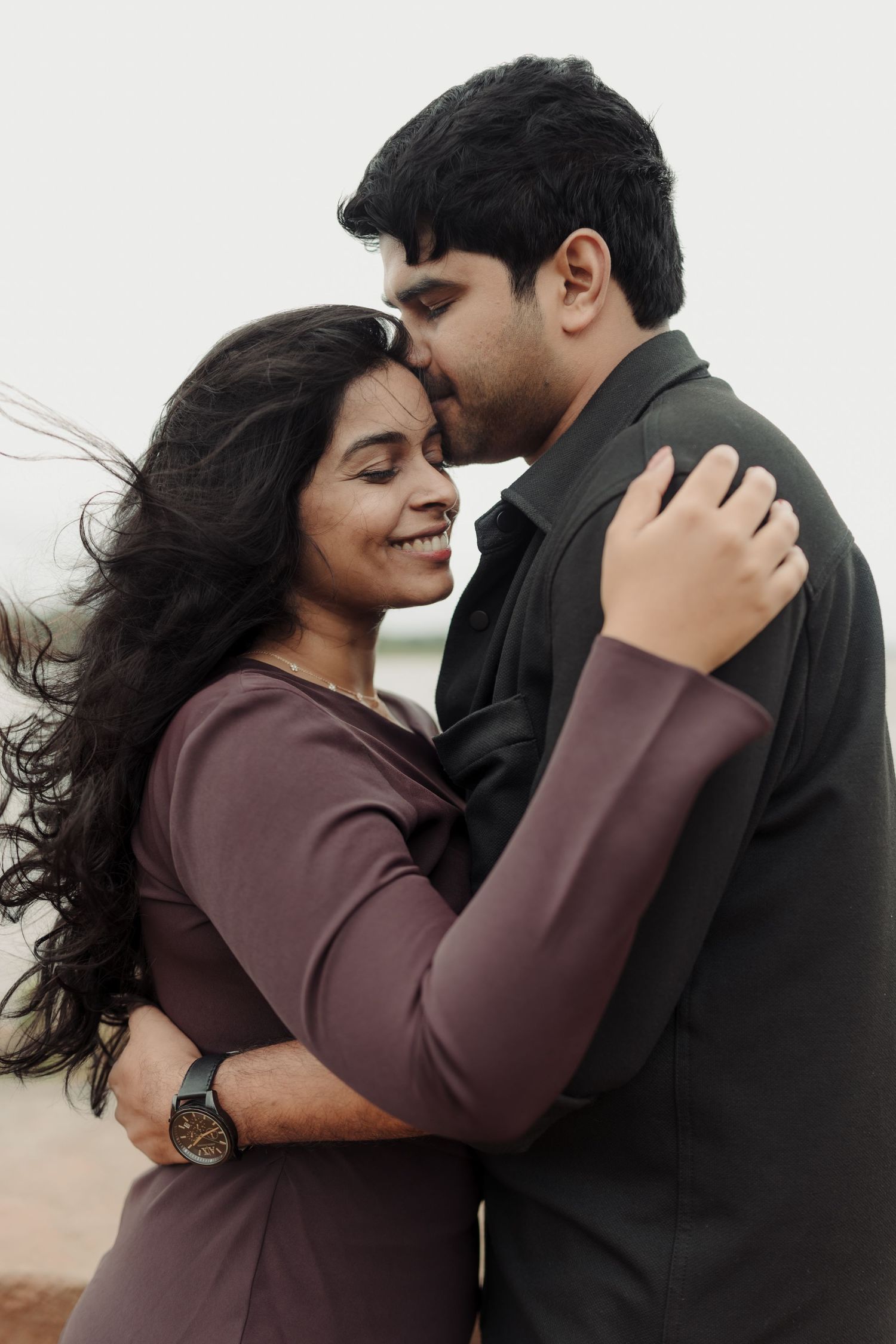 A romantic couple in maroon and black outfits embrace while smiling outdoors during a sunset.
