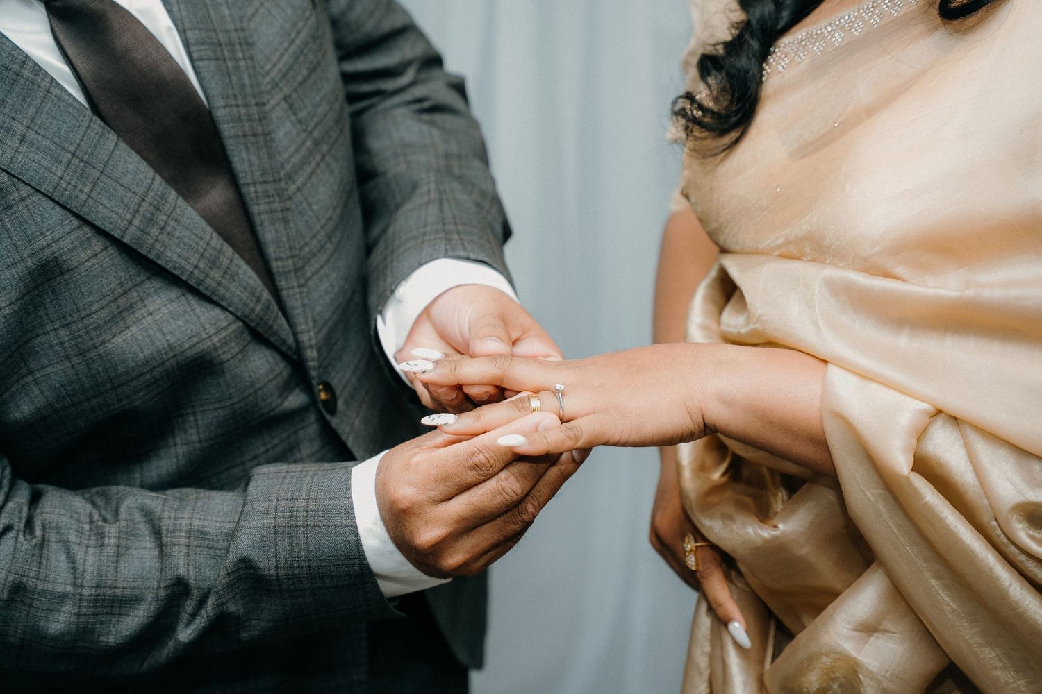 Close-up shot of hands exchanging wedding rings during an elegant ceremony with grey suit and peach dress visible.