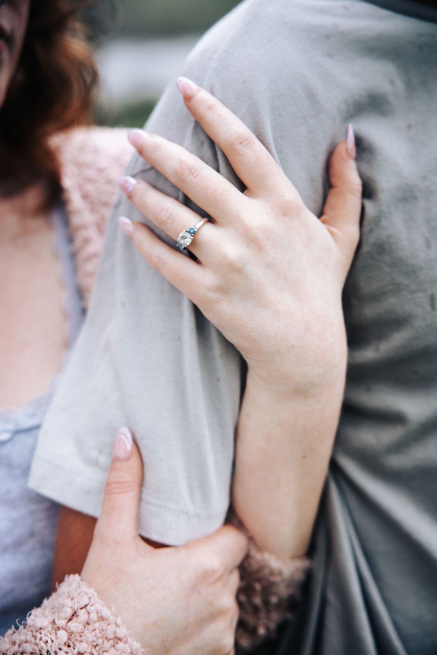 Close-up of a delicate hand wearing an engagement ring resting against gray fabric with pink lace details.