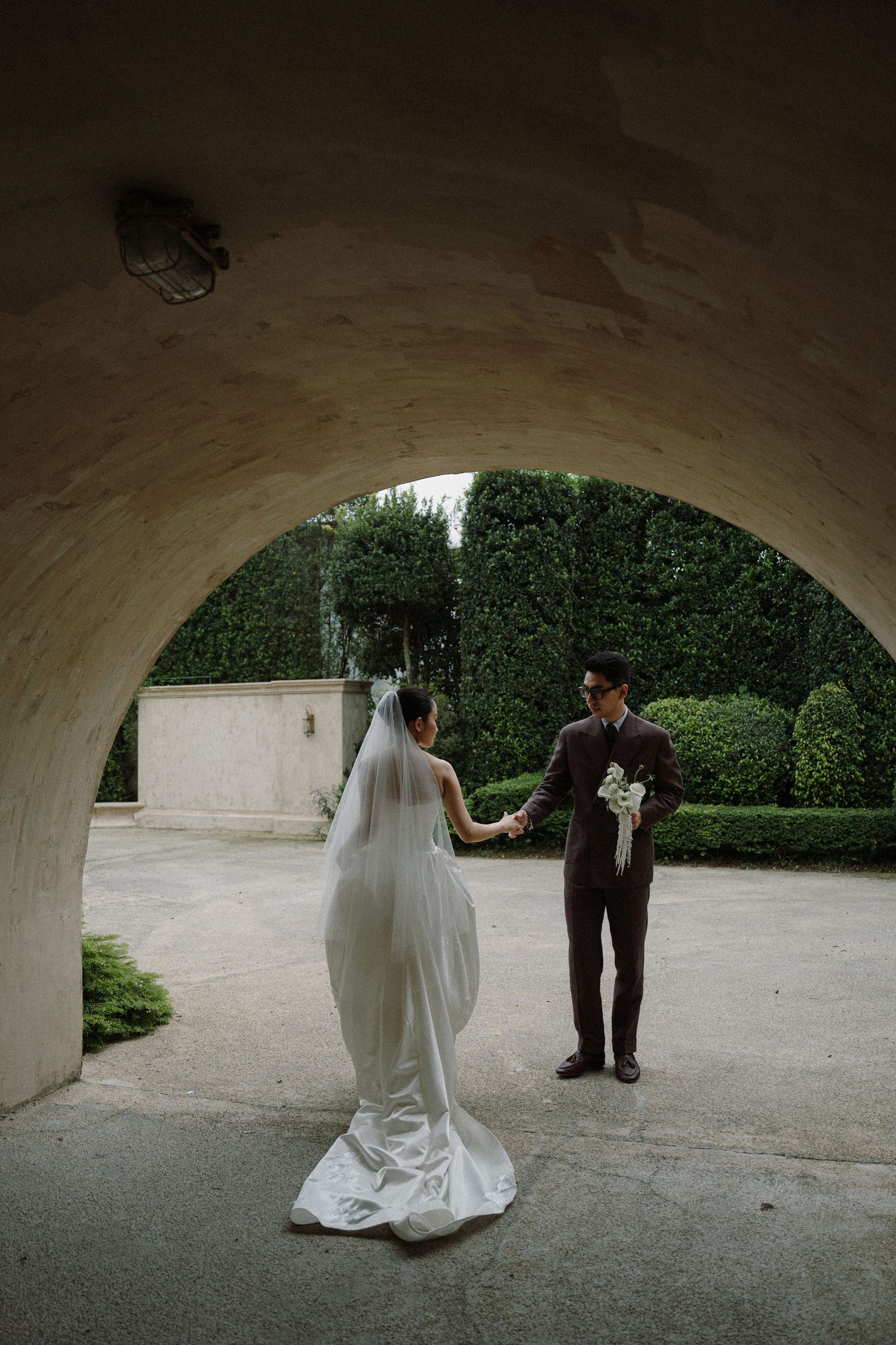 Wedding couple poses under stone archway in garden setting with dramatic lighting.